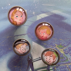 May include: Four round glass cabochons with silver metal frames, each featuring a different character from the movie Peter Pan. The characters are Wendy Darling, John Darling, Peter Pan, and Captain Hook.