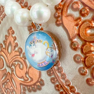 May include: A circular pendant with a cartoon image of Mrs. Potts and Chip from Beauty and the Beast, set against a light blue backdrop. The pendant is attached to a strand of large, white, pearl-like beads. The background has a copper-coloured, ornate design.