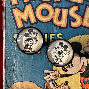 May include: Two round, black-rimmed buttons featuring Mickey Mouse in black and white. The buttons are set against a vintage blue and yellow background with the word "MOUSE" visible. The buttons are likely for clothing or crafts.