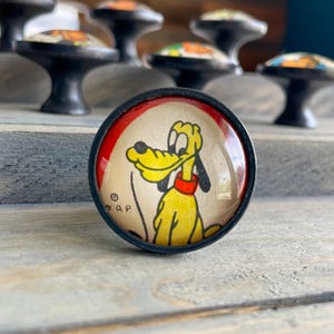 May include: A round cabinet knob with a black base and a domed glass top featuring a yellow cartoon dog with a red collar. The illustration is set against a cream background with a red border and includes the copyright symbol and letters D P.