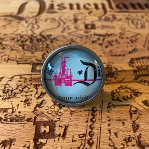 May include: A silver-toned, round, glass-topped knob with a pink and white image of Cinderella's castle and the letter "D" in black script. The text "your visit" is visible at the bottom of the knob.
