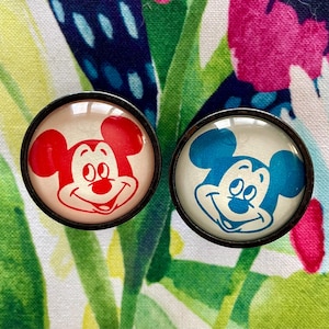 May include: Two round, white, glass Mickey Mouse buttons with black outlines. One button has red ears and the other has blue ears.