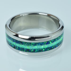 Könnte beinhalten: Ein silberner Ring mit breitem Band. Der Ring hat einen leuchtenden grünen und blauen Opal-Inlay, das das Band umschließt. Der Opal hat ein schimmerndes, gesprenkeltes Aussehen. Der Ring ist vor einem weißen Hintergrund platziert.