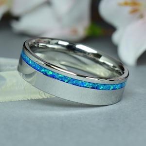 Puede incluir: Un anillo plateado con una banda de ópalo azul vibrante. El anillo se encuentra sobre un fondo gris suave, con flores blancas en el fondo. La banda de ópalo añade un toque de color a la plata pulida.