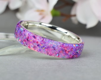May include: A silver band ring with a purple and pink opal inlay. The ring is set against a gray background with blurred pink flowers in the background.