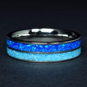 Puede incluir: Un anillo plateado con dos bandas de piedras incrustadas azules y celeste. La banda superior es de un azul vibrante, mientras que la banda inferior es de un color turquesa más claro. El anillo está sobre un fondo oscuro.
