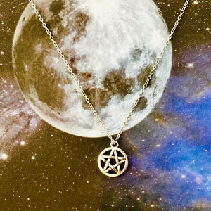 Puede incluir: Un collar de cadena de plata con un colgante de pentagrama. El collar está fotografiado sobre un fondo de luna y estrellas.