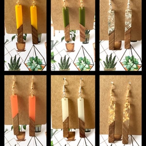 May include: Six pairs of handmade resin and wood earrings. Each pair features a rectangular resin piece with a different colour and pattern, including yellow, green, clear with gold flakes, orange, white, and gold flakes. The resin pieces are attached to a wooden base.