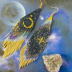 Puede incluir: Un par de aretes largos de cuentas con un tema celestial. Los aretes son negros con detalles dorados y plateados, y presentan una luna creciente y estrellas.