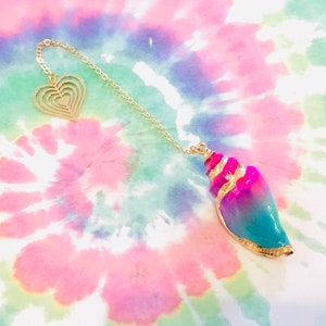 K&ouml;nnte beinhalten: Eine goldene Kette mit einem herzf&ouml;rmigen Anh&auml;nger und einem Muschel-Anh&auml;nger. Die Muschel ist rosa, blau und gr&uuml;n mit goldenen Verzierungen.