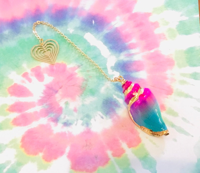 K&ouml;nnte beinhalten: Eine goldene Kette mit einem herzf&ouml;rmigen Anh&auml;nger und einem Muschel-Anh&auml;nger. Die Muschel ist rosa, blau und gr&uuml;n mit goldenen Verzierungen.