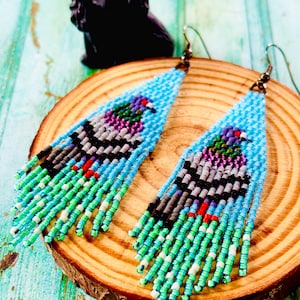 May include: A pair of beaded earrings with a bird design. The earrings have a light blue background with a bird design in shades of purple, grey, black, and green. The fringe is turquoise and green. The earrings are displayed on a wooden surface.