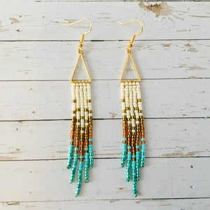 May include: Beaded earrings with gold-coloured triangle tops and ear wires. The earrings have a gradient design, transitioning from cream to gold, copper, and turquoise beads. The earrings are displayed against a white wooden background.