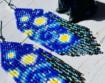 May include: A pair of long, blue and white beaded earrings with a starry night design. The earrings are made with small, colorful beads and have a fringe-like bottom.