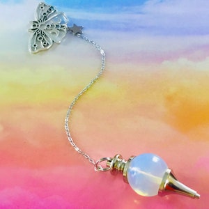 Puede incluir: Una cadena de plata con un dije de mariposa y un dije de estrella unidos a un péndulo de ópalo blanco. El péndulo es una bola redonda con una punta.