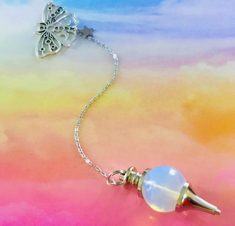 K&ouml;nnte beinhalten: Eine silberne Kette mit einem Schmetterlingsanh&auml;nger und einem Sternanh&auml;nger, die an einem wei&szlig;en Opal-Pendel befestigt sind. Das Pendel ist eine runde Kugel mit einer Spitze.