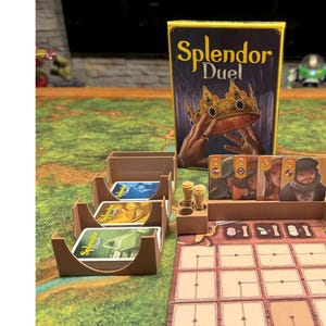 Może przedstawiać: Drewniany organizer do gier w kolorze brązowym z miejscami na karty i planszą do gry planszowej "Splendor Duel". Pudełko gry jest widoczne w tle ze złotą koroną i tytułem gry.