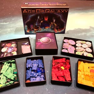 Może przedstawiać: Gra planszowa o nazwie "Age of Galaxy" z pudełkiem i różnymi komponentami, w tym planszą do gry, kartami, żetonami i kostkami. Plansza do gry jest fioletowa i przedstawia galaktykę. Komponenty są zorganizowane w oddzielnych tacach.