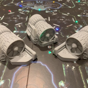 Peut inclure: Trois pièces de jeu grises imprimées en 3D, ressemblant à des moteurs à réaction, sont disposées sur un plateau de jeu. Le plateau a un fond sombre avec un design futuriste et des éléments lumineux. Les pièces sont étiquetées "Д-1" et "Д-3".