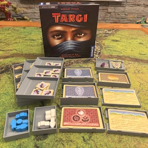Op de afbeelding: Een bordspel genaamd "Targi: Ancient Tribes" met een doos met een afbeelding van een vrouw met een zwarte hoofddoek. Het spel bevat verschillende kaarten, tokens en trays.