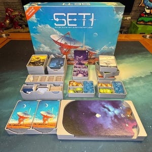 Könnte beinhalten: Ein Spielorganizer für das Brettspiel SETI. Der Organizer ist aus grauem Kunststoff und hat Fächer für die Karten, Spielsteine und andere Komponenten des Spiels. Die Spielbox befindet sich im Hintergrund und hat ein blaues und weißes Design mit dem Spieltitel "SETI", der in weiß gedruckt ist. Die Spielbox zeigt auch ein Bild eines großen Radioteleskops.