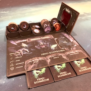 Könnte beinhalten: Ein braunes Holzspielbrett mit Fächern für Spielsteine und Karten. Das Brett hat ein gedrucktes Design mit dem Text "XENO-TAFEL" und "XENO-SCHWÄCHEN".