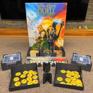Könnte beinhalten: Eine schwarz-weiße Spielbox mit dem Text "Der Herr der Ringe Duell Mittelerde" auf der Vorderseite. Die Box ist geöffnet und zeigt die Spielkomponenten, darunter ein schwarz-weißes Spielbrett, schwarz-weiße Spielfiguren und eine schwarz-weiße Spielkarte.