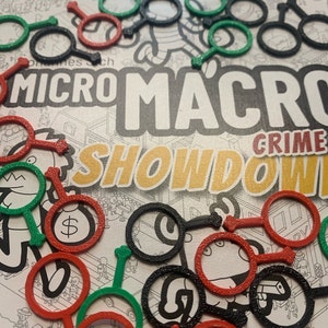 May include: A close-up of a game board with black, red and green game pieces. The game board has the text "Micro Macro Crime Showdown" printed on it.