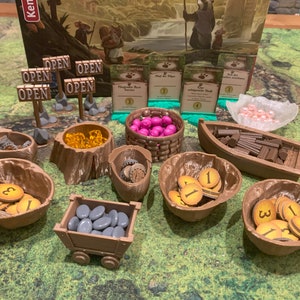 Könnte beinhalten: Ein Spielbrett mit verschiedenen Holzmarkern und Behältern. Die Marker sind in verschiedenen Farben, darunter Gold, Pink und Braun. Die Behälter sind aus Holz und haben verschiedene Formen, darunter Schalen, Körbe und ein kleiner Wagen. Das Spielbrett ist aus grünem Material und hat einen braunen Rand.