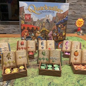Op de afbeelding: Een bordspel genaamd "De Quacksalber van Quedlinburg" met een kleurrijke doos en spelstukken. Het spel speelt zich af in een middeleeuwse stad met een marktplein en verschillende winkels. De spelstukken omvatten een verscheidenheid aan kleurrijke tokens, dobbelstenen en kaarten.