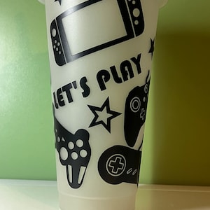 May include: A clear plastic tumbler with a lid, decorated with black gaming controller graphics, stars, and a game console. The words "LET'S PLAY" are printed in black. The tumbler is designed for cold drinks.