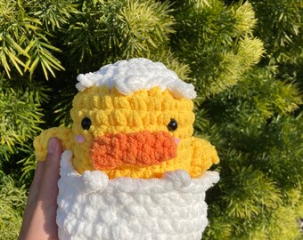 Puede incluir: Un patito amarillo de crochet asomando de un huevo blanco de crochet. El patito tiene un pequeño pico naranja y ojos negros.