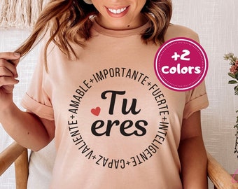 Op de afbeelding: Een lichtroze T-shirt met een zwarte cirkelvormige afbeelding waarop "Tu eres" staat met een klein rood hartje. De cirkelvormige afbeelding is omringd door de woorden "Valiente", "Capaz", "Inteligente", "Fuerte", "Importante" en "Amable". Het shirt is verkrijgbaar in twee kleuren.