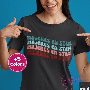 May include: Black t-shirt with the text "MUJERES EN STEM" in a retro font. The text is stacked in multiple rows with teal, coral, and red colors. The image also shows a "+5 colors" button and the word "BLACK".