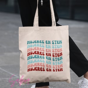 May include: A beige tote bag with the text "MUJERES EN STEM" repeated in a colorful, wavy font.