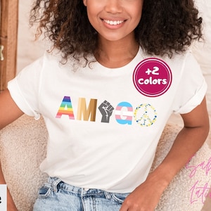 May include: White t-shirt with the word "AMIGO" spelled out in colorful letters and symbols, including a rainbow, a fist, a transgender symbol, and a peace sign. The shirt also has a pink circle with the text " +2 Colors" on it.