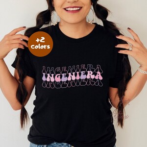 May include: Black t-shirt with the word "INGENIERA" in a retro, wavy font in shades of pink and purple. The shirt has a crew neck and short sleeves. A brown circle with the text "+2 colors" is in the upper left corner.