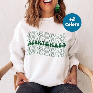 May include: White sweatshirt featuring the word "AFORTUNADA" in a green and white wavy design. The text is repeated across the front. The sweatshirt has a crew neck and long sleeves, suitable for cooler weather.