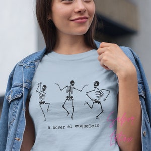 May include: Light blue t-shirt featuring three dancing skeleton figures in black. The phrase "a mover el esqueleto" is printed below the skeletons. The shirt is a baby blue color.