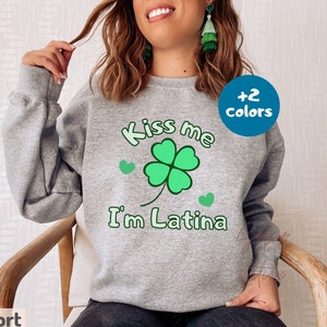 May include: A light gray sweatshirt with the text "Kiss me I'm Latina" in green, along with a green four-leaf clover and two green hearts. The sweatshirt has a crew neck and long sleeves.
