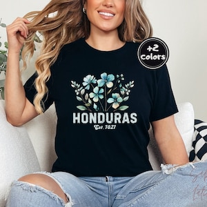 May include: A black t-shirt with a floral design and the text "HONDURAS Est. 1827". The model is wearing blue jeans and sitting on a white couch.