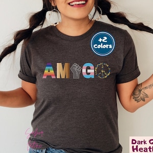May include: A dark grey heather t-shirt with the text "AMIGO" in rainbow colors, a black fist, a transgender pride flag, and a peace sign. The text "+2 colors" is on a blue circle.