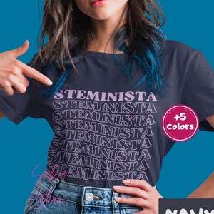 May include: A navy blue t-shirt with the word "STEMINISTA" repeated in a stacked pattern in a light purple color. The shirt is shown on a model with blue hair. A pink circle with the text " +5 colors" is in the upper right corner of the image. The text "NAVY" is in the lower right corner of the image.