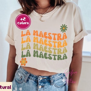 May include: A cream-colored t-shirt with the text "LA MAESTRA" in orange, yellow, and green, with flower graphics. The shirt has rolled-up sleeves and is paired with blue jeans. The text "+2 colors" is in a pink circle.