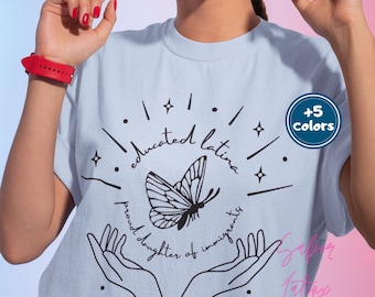 May include: A light blue t-shirt with a butterfly graphic and the text "educated latina proud daughter of immigrants". The shirt is available in 5 colors.
