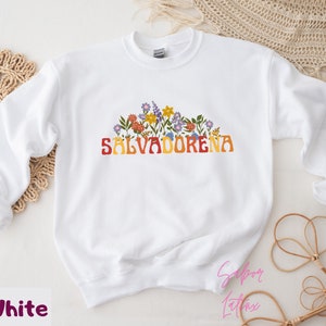 May include: White sweatshirt with a floral design and the text "SALVADORENA" in a retro font.