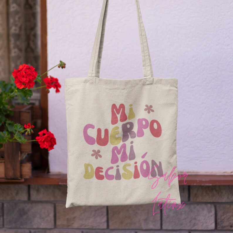 May include: A beige tote bag with the text "Mi Cuerpo Mi Decisi&oacute;n" in colorful, bubble-style letters. The bag has two handles and is made of a canvas-like material. The design includes small flower graphics.