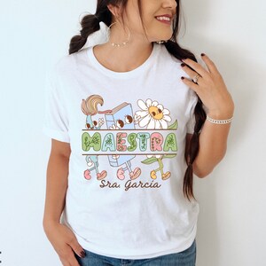 Peut inclure: T-shirt blanc avec un motif graphique color&eacute; repr&eacute;sentant un livre, un pinceau et des fleurs. Le texte "MAESTRA Sra. Garcia" est imprim&eacute; sur le t-shirt.