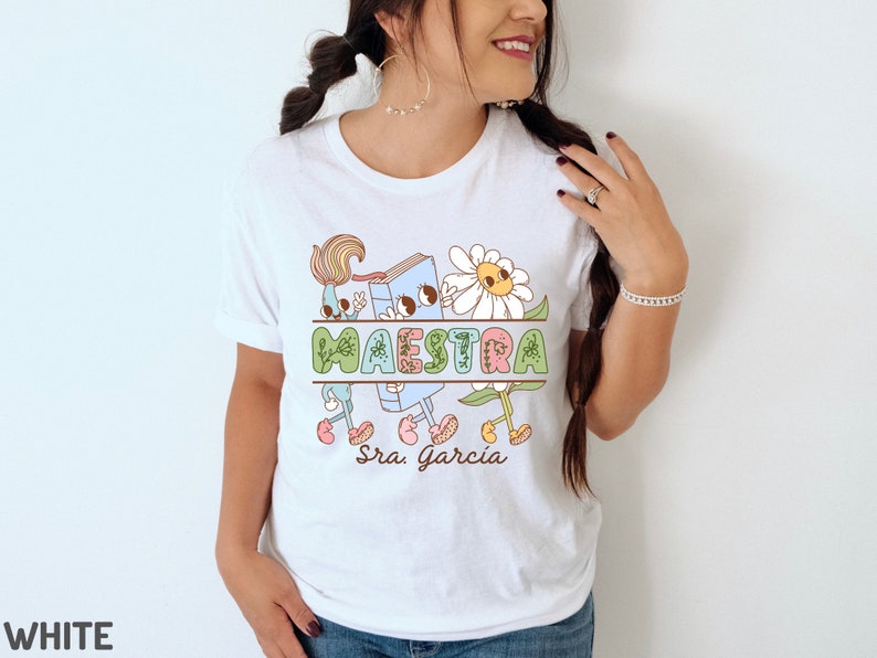 Peut inclure: T-shirt blanc avec un motif graphique color&eacute; repr&eacute;sentant un livre, un pinceau et des fleurs. Le texte "MAESTRA Sra. Garcia" est imprim&eacute; sur le t-shirt.
