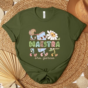 Peut inclure: T-shirt vert olive avec une illustration de dessin anim&eacute; d'un livre, d'une fleur et du mot "MAESTRA" avec le texte "Sra. Garcia" en dessous.